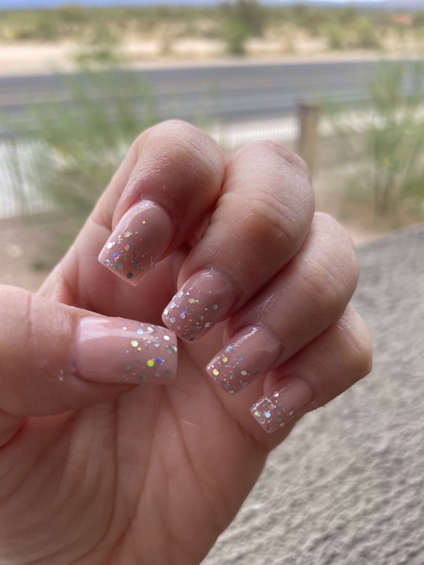 Clear nude coffin-shaped nails with a light scatter of glitter, a desert roadside behind them