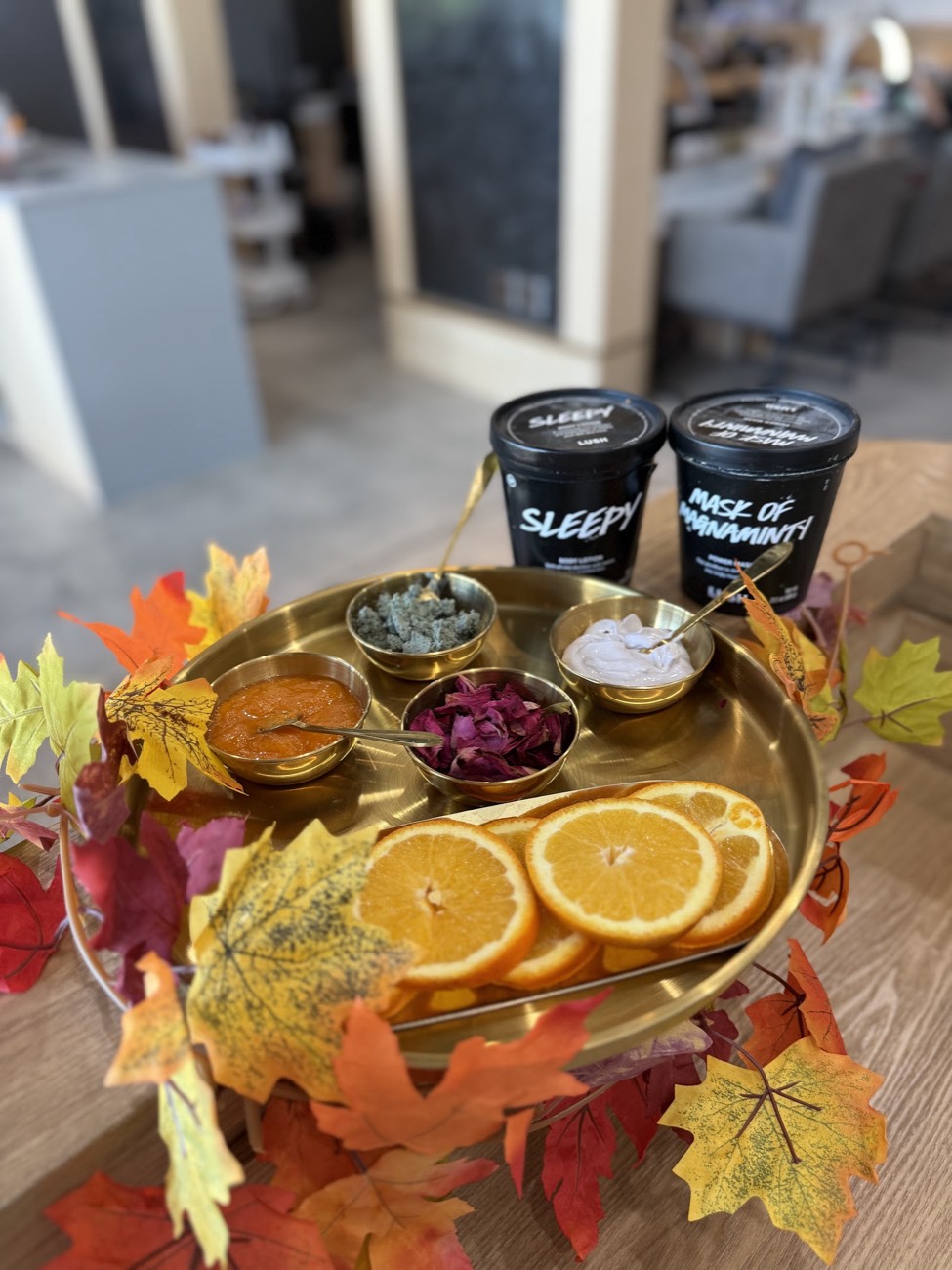 A gold tray with fresh orange slices, small dishes of scrub and mask and two Lush body-care tubs, framed by autumn leaves on a wooden table